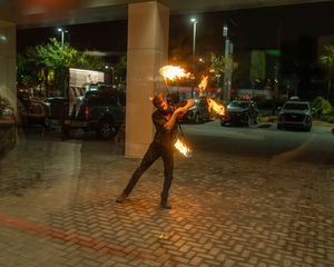 Fire performer spins flaming batons in a dramatic nighttime display on a brick patio.