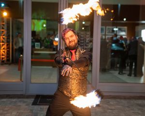 Fire artist in metallic vest performs with multiple flames against illuminated storefront.
