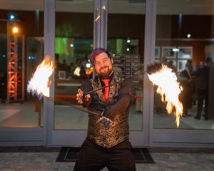 Fire dancer performs with flaming props against illuminated building columns at night.