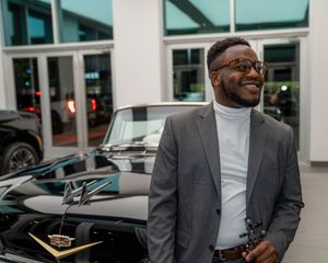 Professional in a gray suit and white shirt stands near parked cars while holding documents.
