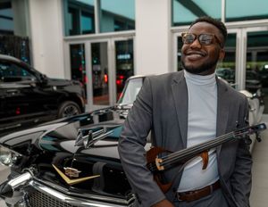 Professional in a grey blazer and turtleneck smiles while standing next to a car with a violin.