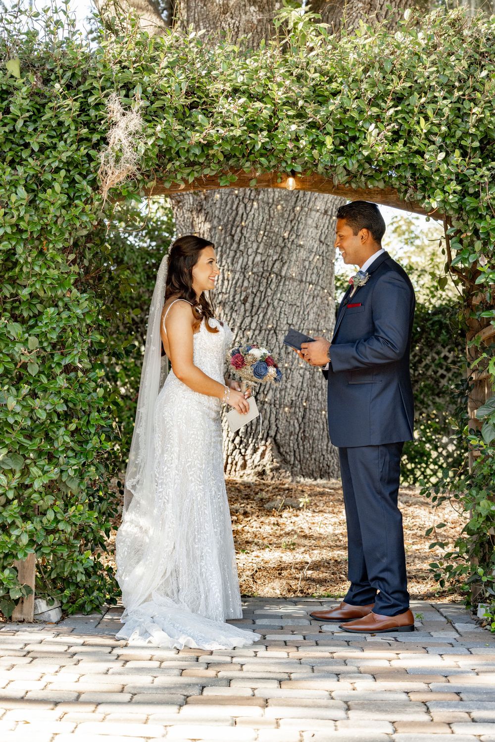 A bride and groom share a romantic moment on a brick pathway surrounded by lush greenery and trees.