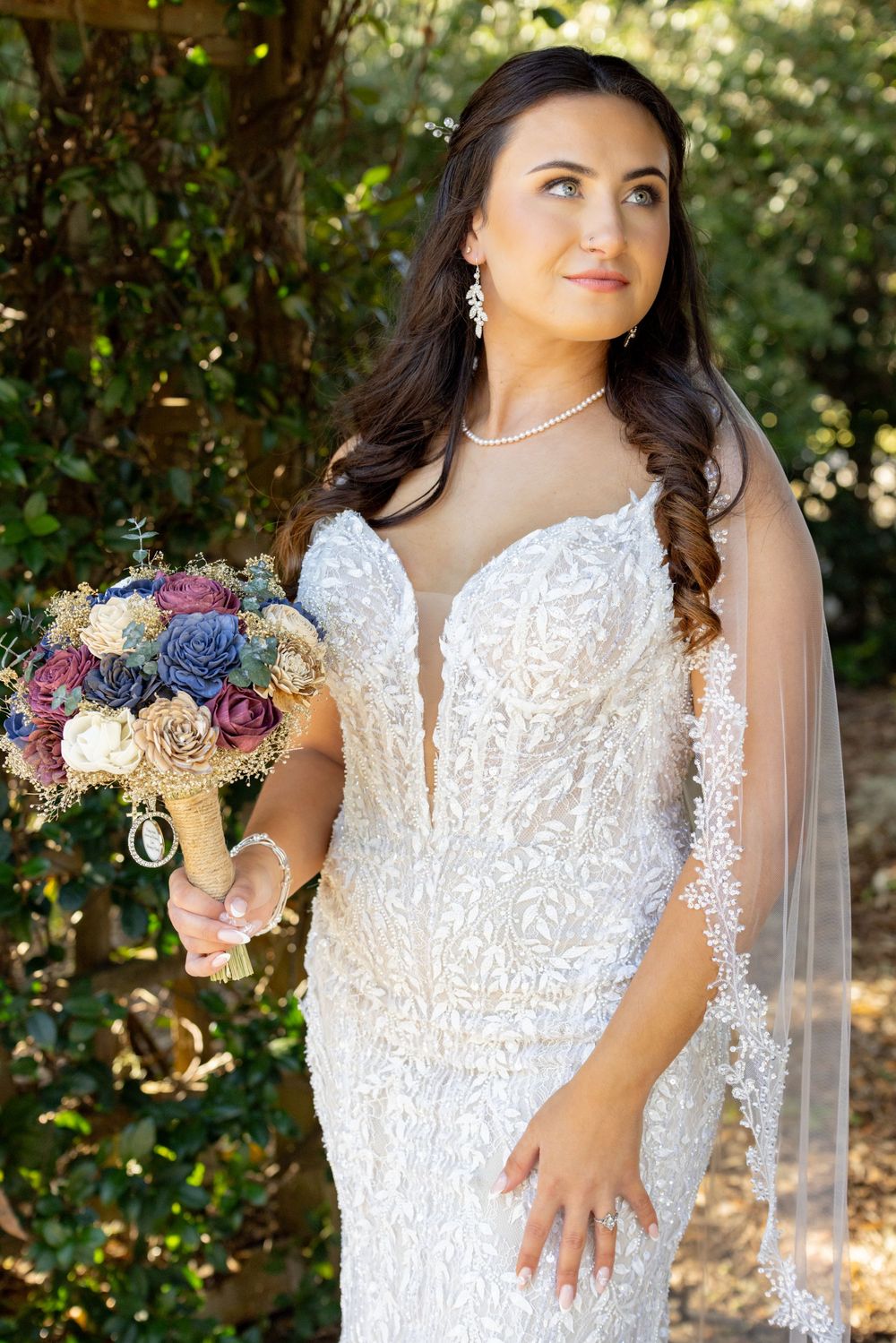 Bride in detailed lace wedding dress poses with bouquet against natural garden backdrop.