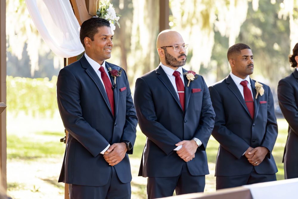 A line of groomsmen in matching navy suits with red ties stand outdoors during a wedding ceremony.