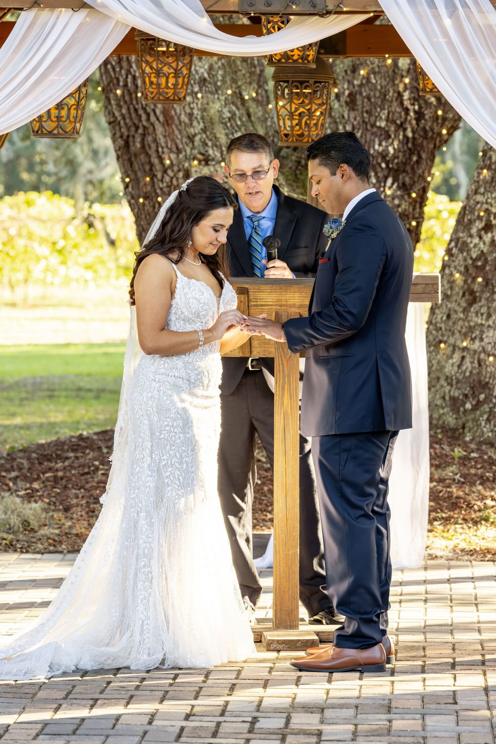 Wedding ceremony under white draped gazebo with brick paved flooring and trees in background.