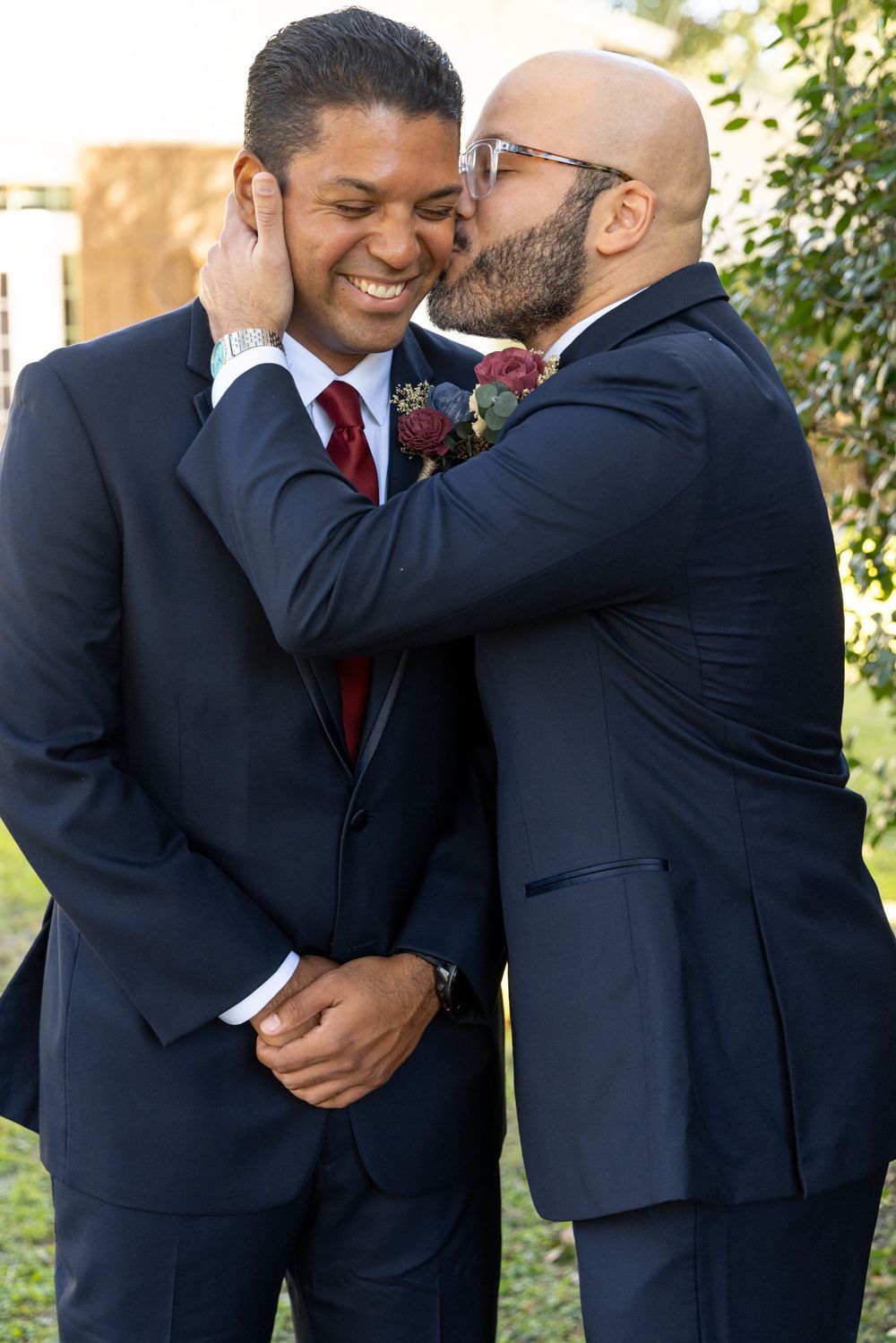 Two people in dark suits share an intimate moment outdoors during a wedding photoshoot.