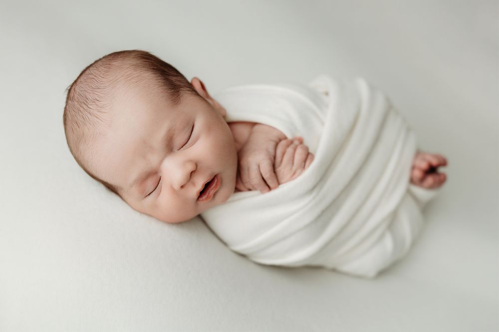 A sleeping newborn baby wrapped in soft white fabric is photographed in a collage of peaceful poses.