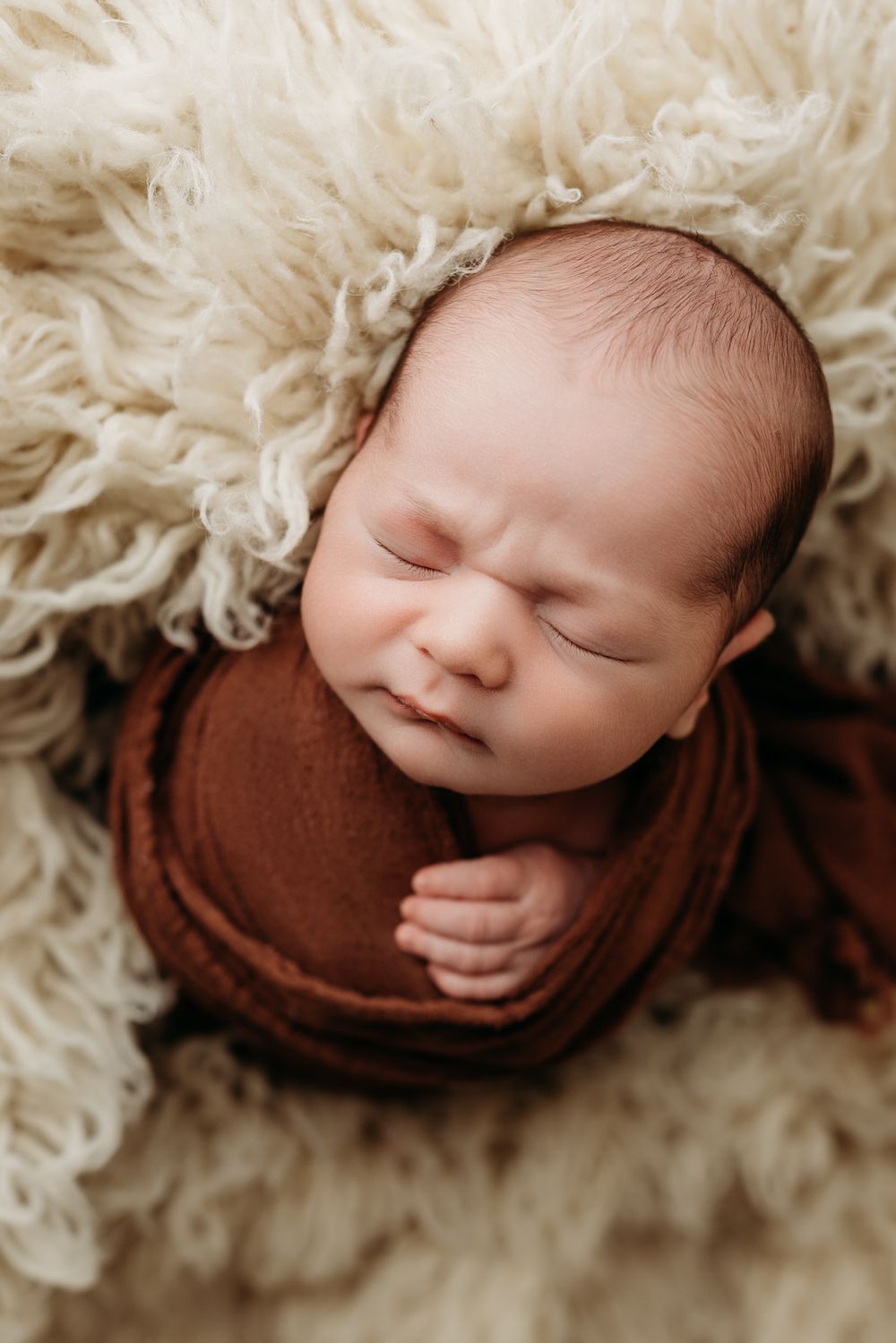 Peaceful sleeping newborn wrapped in brown blanket nestled in soft white fur during professional photo session.