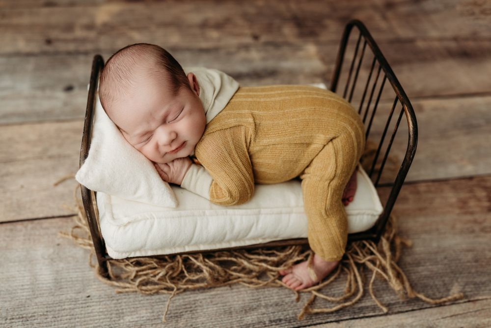 A sleeping baby in mustard yellow outfit rests on a white pillow in a metal frame on rustic wooden floor.