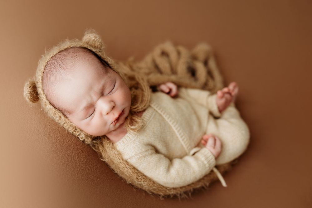 A sleeping newborn baby in cream colored wrap lies peacefully in a woven basket against a brown backdrop.