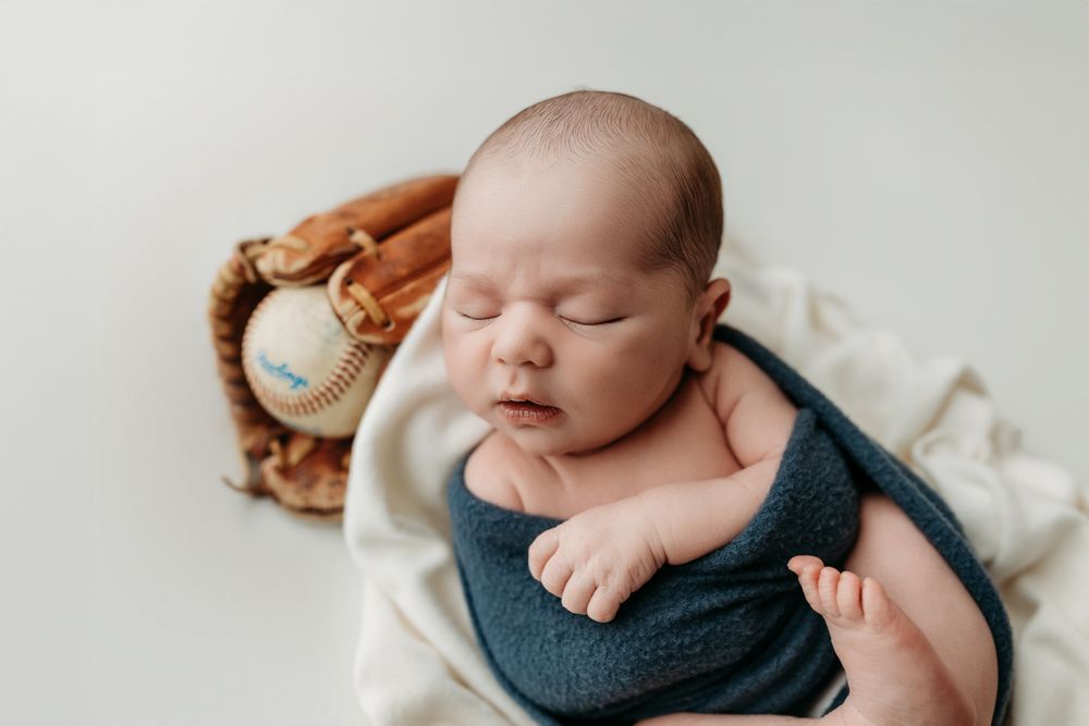 Newborn baby sleeping peacefully on white blanket next to baseball glove and ball.