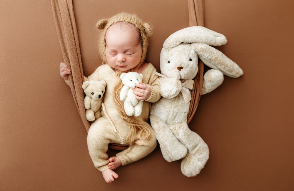 A sleeping baby in beige outfit lies surrounded by plush bunny toys on brown background.