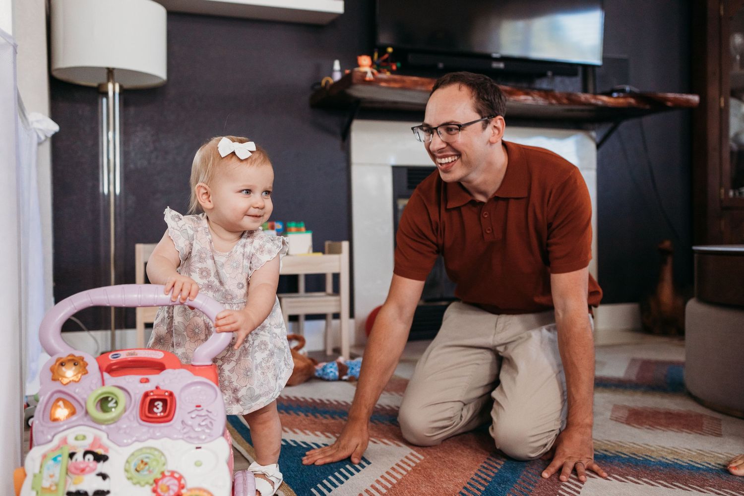 Toddler in a floral dress sits on the floor playing with colorful blocks while adults watch nearby.
