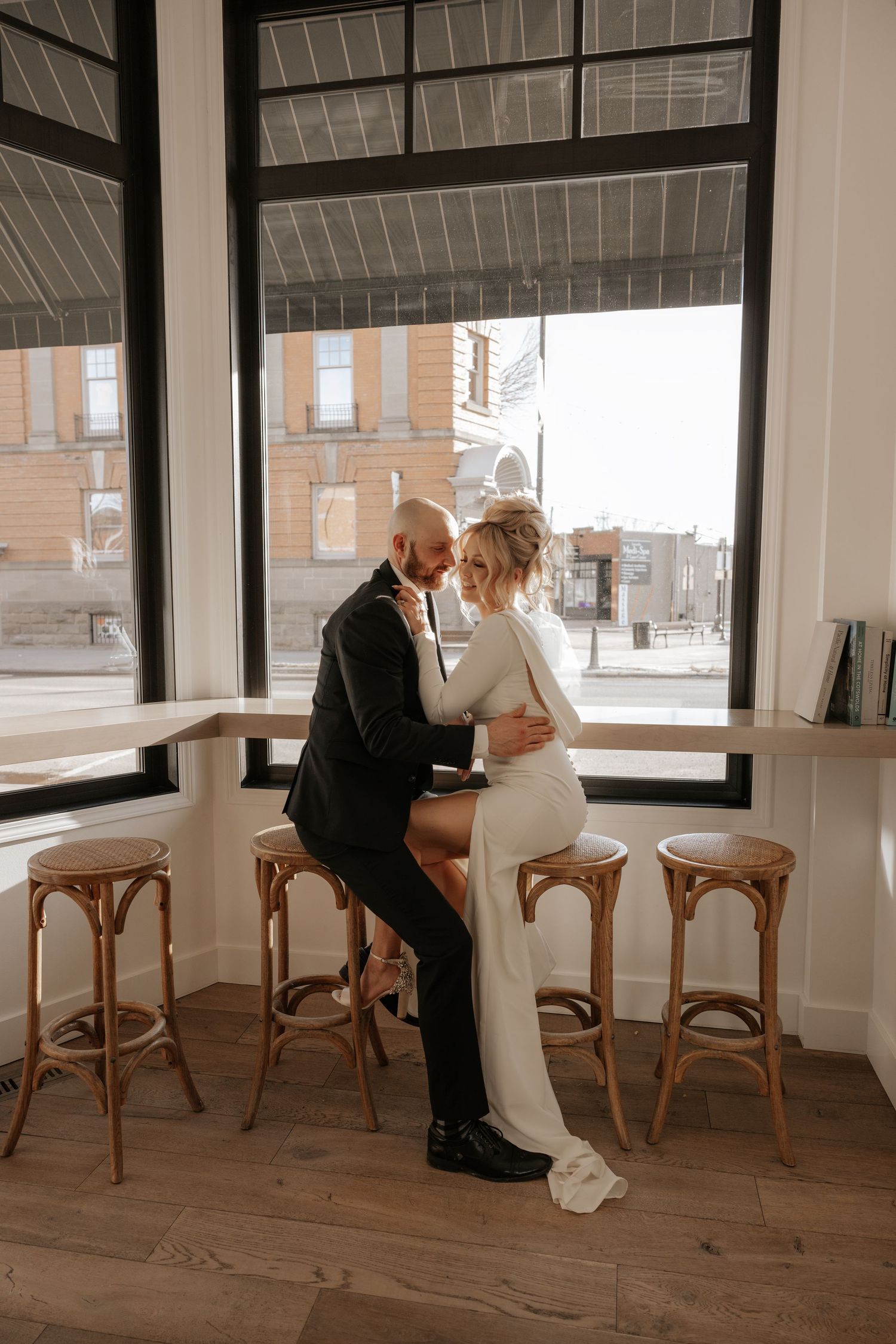 A romantic wedding couple shares an intimate moment in a bright sunlit room with wooden chairs and large windows.