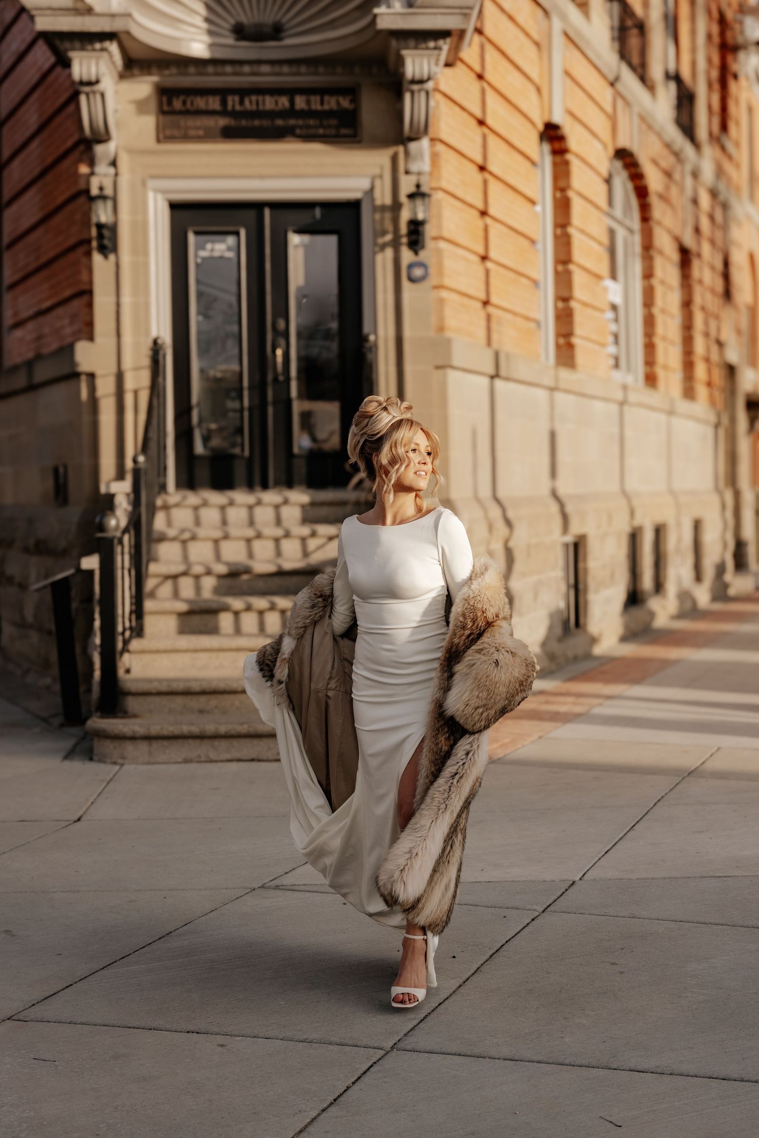 Person in white dress holding teddy bear performs graceful dance movements in front of historic building facade.