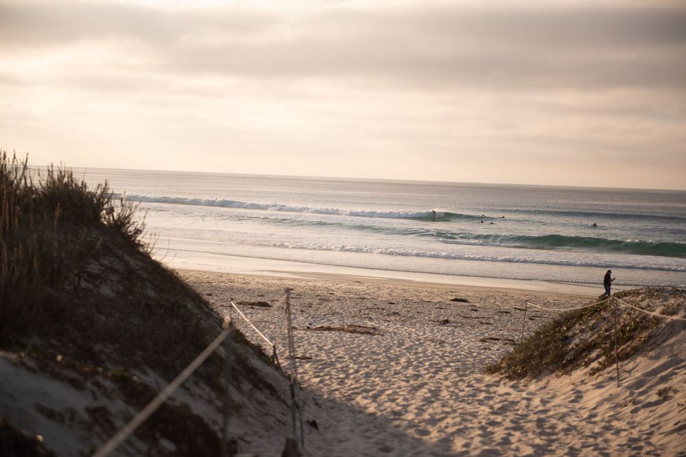 A lone figure walks along a serene beach at sunset with coastal dunes and rolling waves in the background.