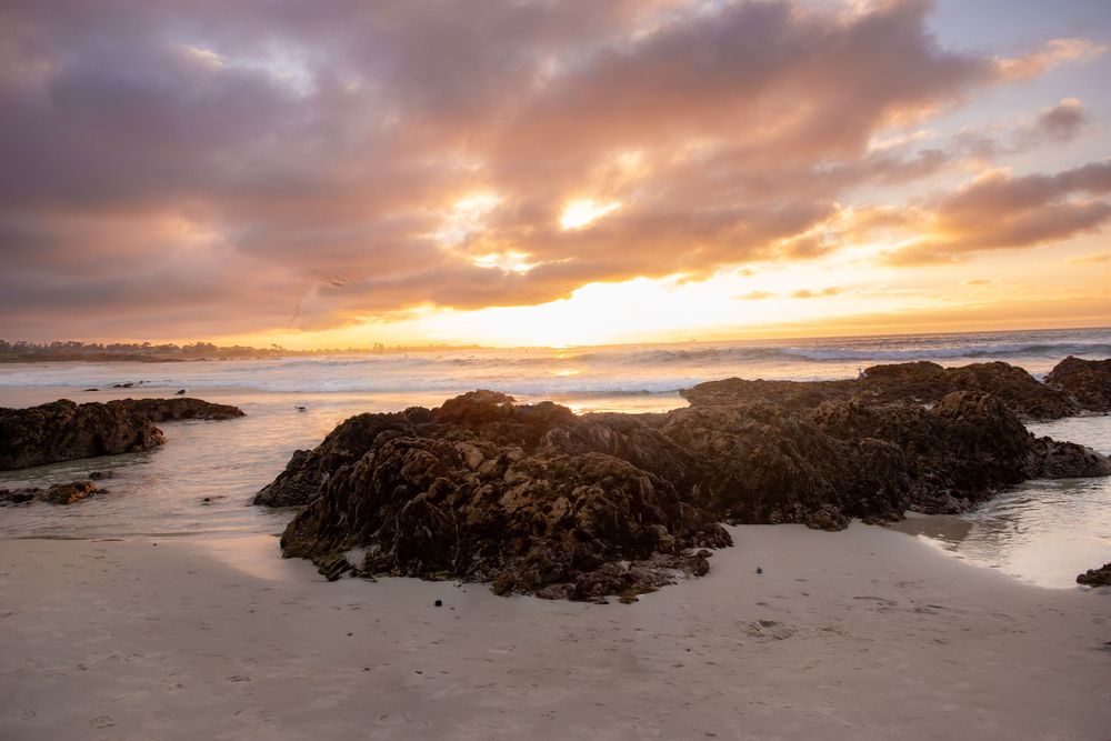 Dramatic sunset illuminates rocky beach landscape with golden light streaming through clouds over coastal waters.