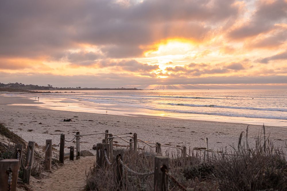Dramatic sunset over a beach with wooden posts and fence remnants silhouetted against an orange and purple sky.