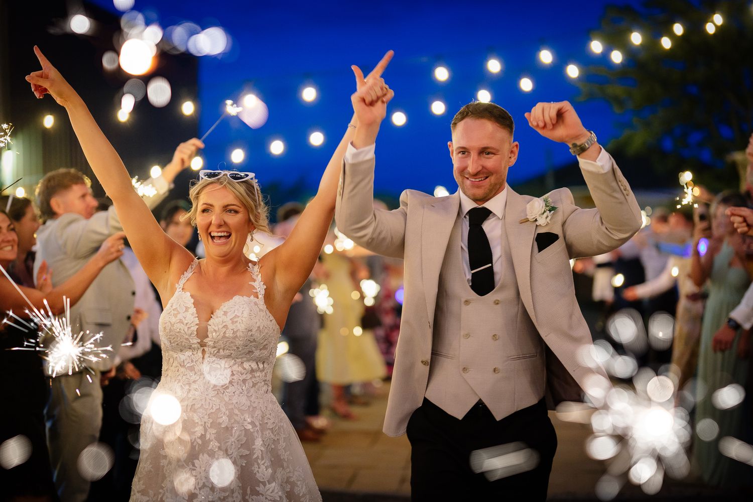 Wedding party celebrates outdoors at night under twinkling string lights and sparklers creating a magical atmosphere.