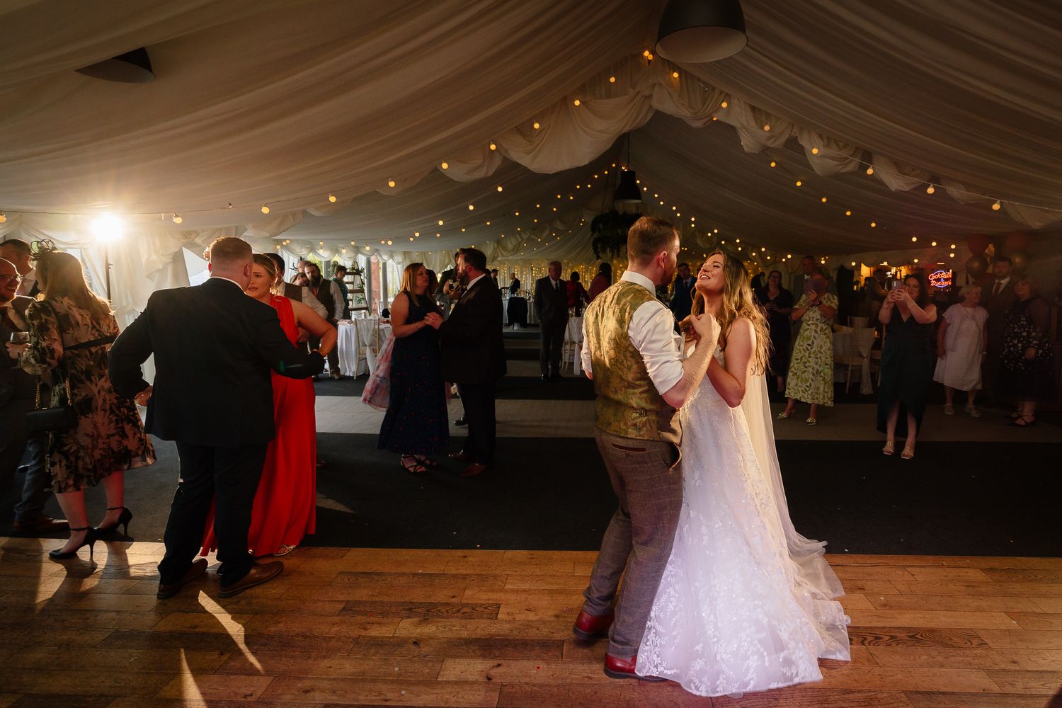 Wedding guests dance together under a tent decorated with twinkling lights during an evening reception celebration.