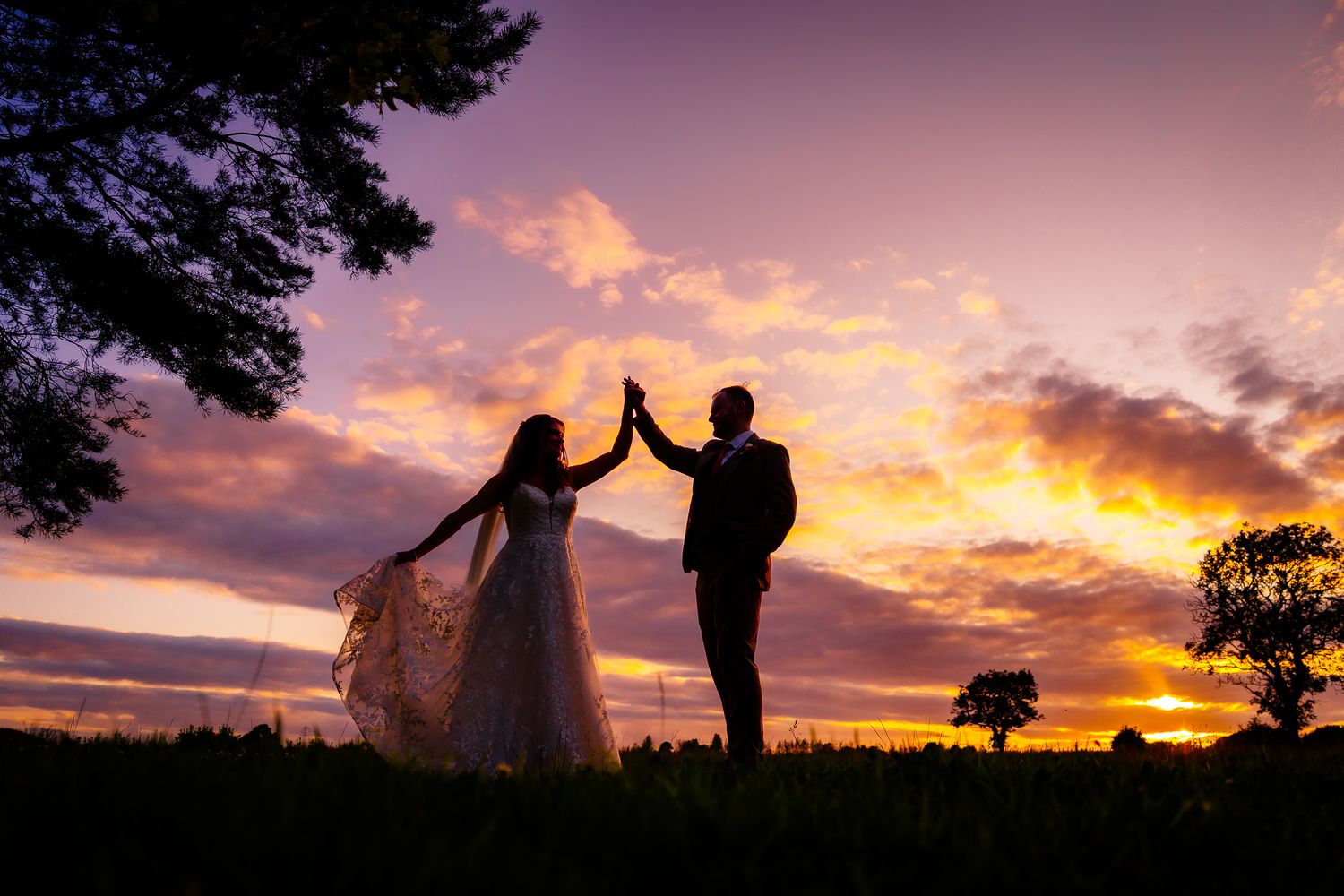 A couple in wedding attire silhouetted against a dramatic purple and orange sunset sky, giving each other a high five.