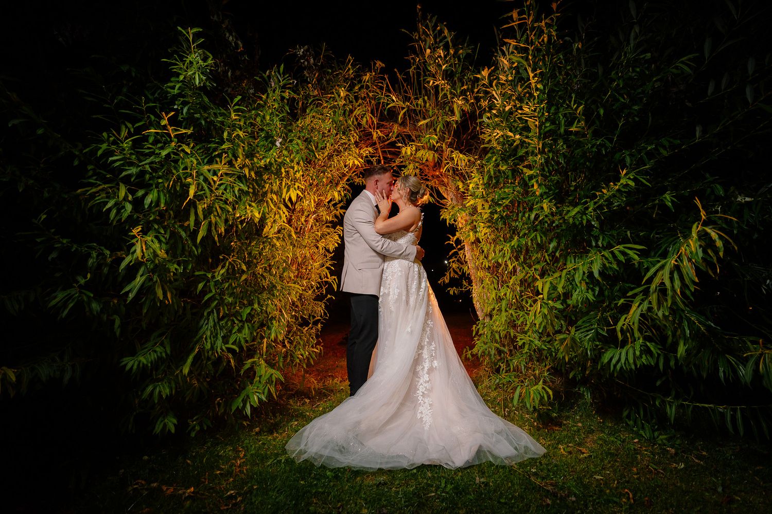 A couple embraces under a glowing natural archway of foliage at night, creating a magical wedding scene.