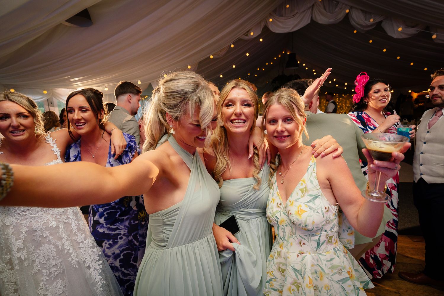 Wedding guests dance and celebrate together on a lively dance floor under twinkling lights at a reception party.