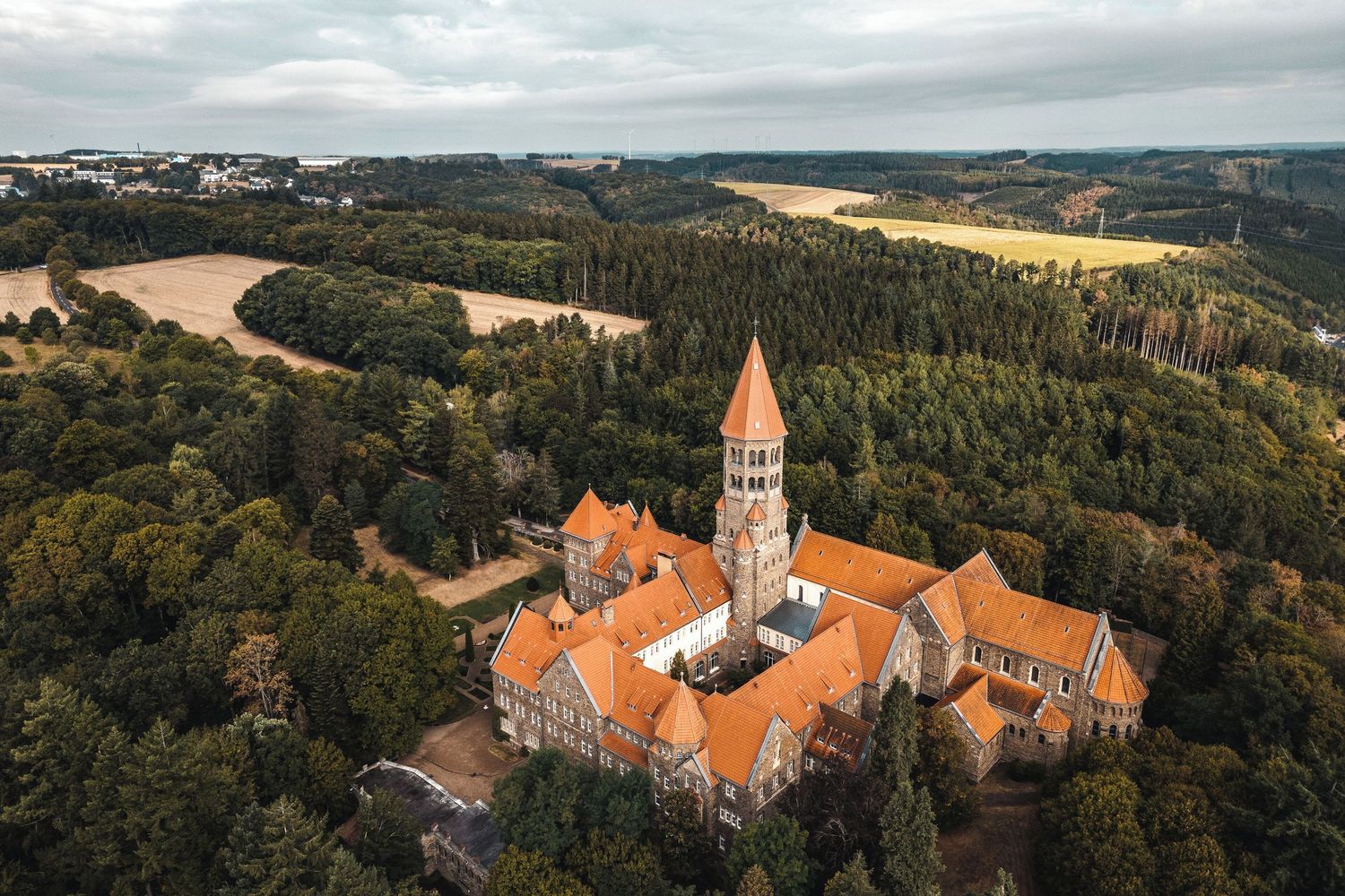 Aerial panoramic view of a historic monastery with orange tiled roofs nestled among forested hills at sunset.