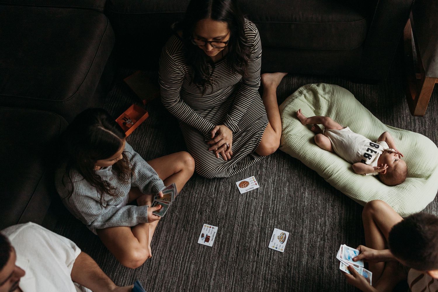 Group of people sitting on floor playing card game with white cards spread out on dark carpet.