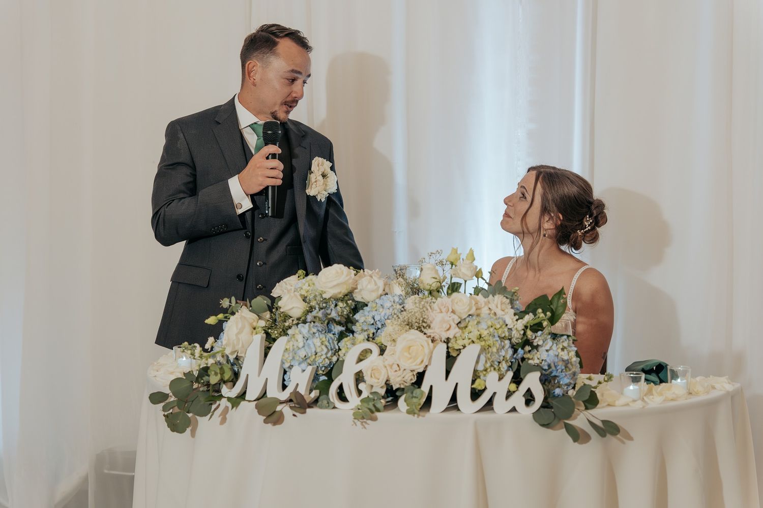 Wedding reception table decorated with white flowers and 'Mr & Mrs' sign while the couple gives a toast to their guests.