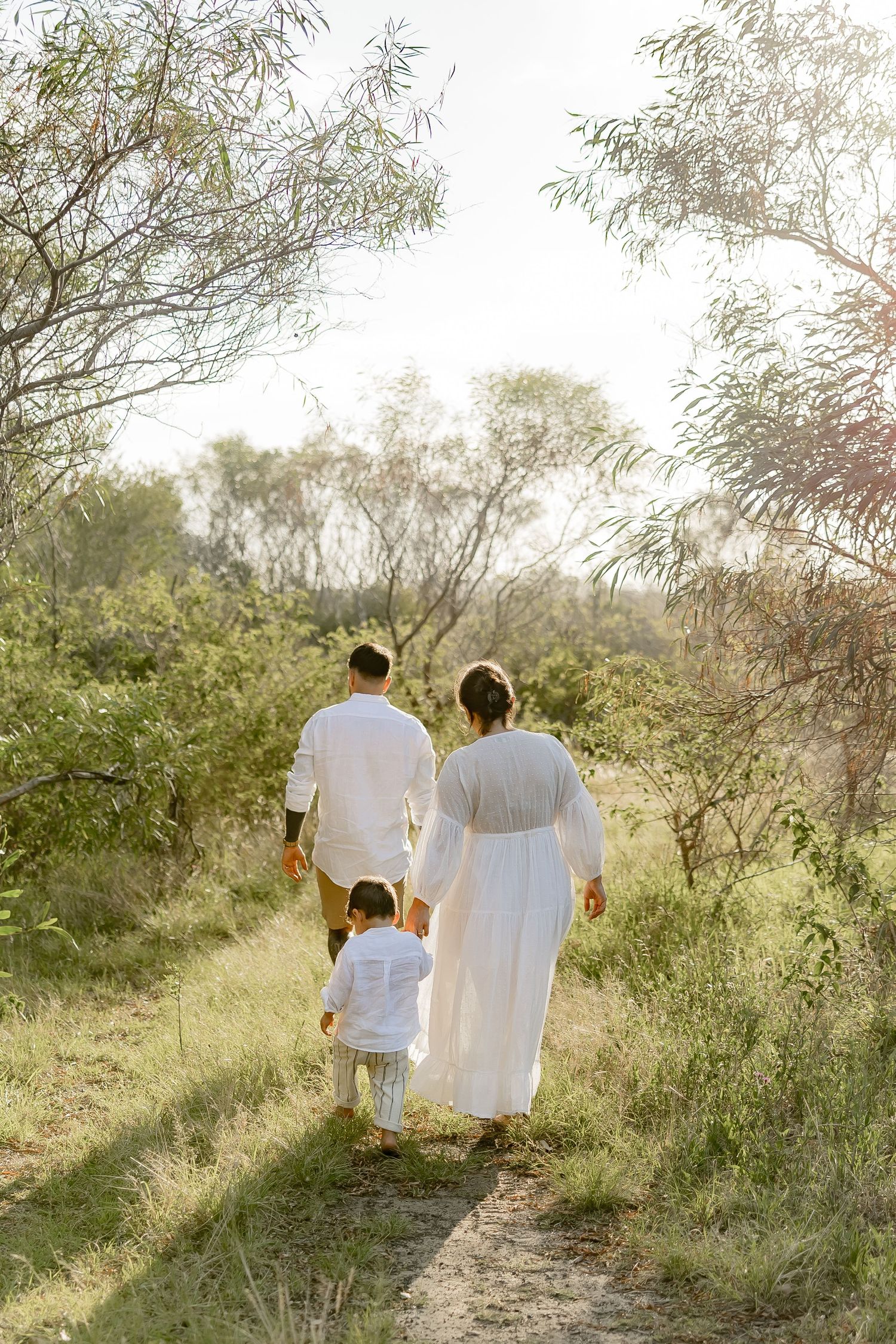 A family in white clothing walks together on a nature trail surrounded by green foliage at sunset.