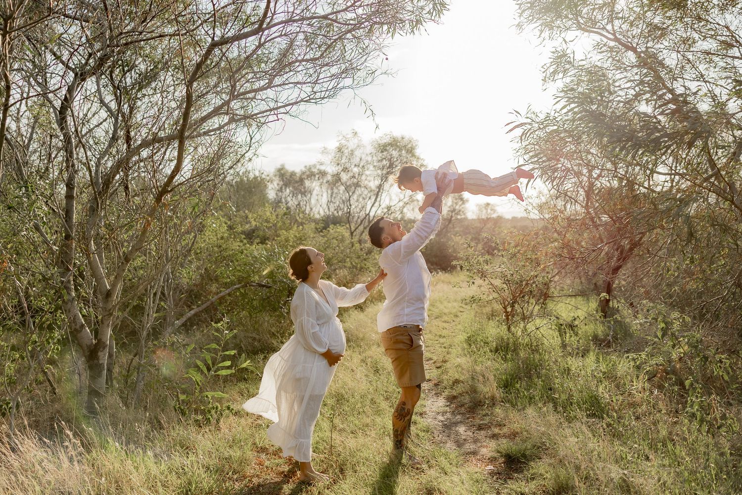 A couple enjoys a playful moment together in a sunlit meadow with trees and bushes.