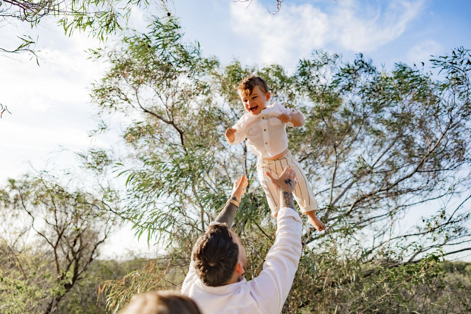 Joyful outdoor family photo session with white outfits and natural sunlight filtering through trees.