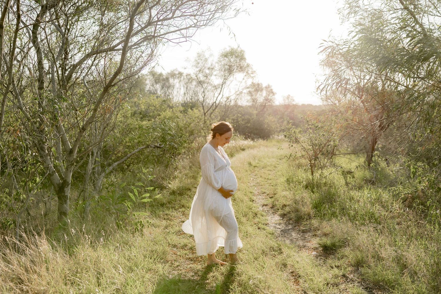 A pregnant woman in a white dress walks through a sunlit field with tall grass and trees.