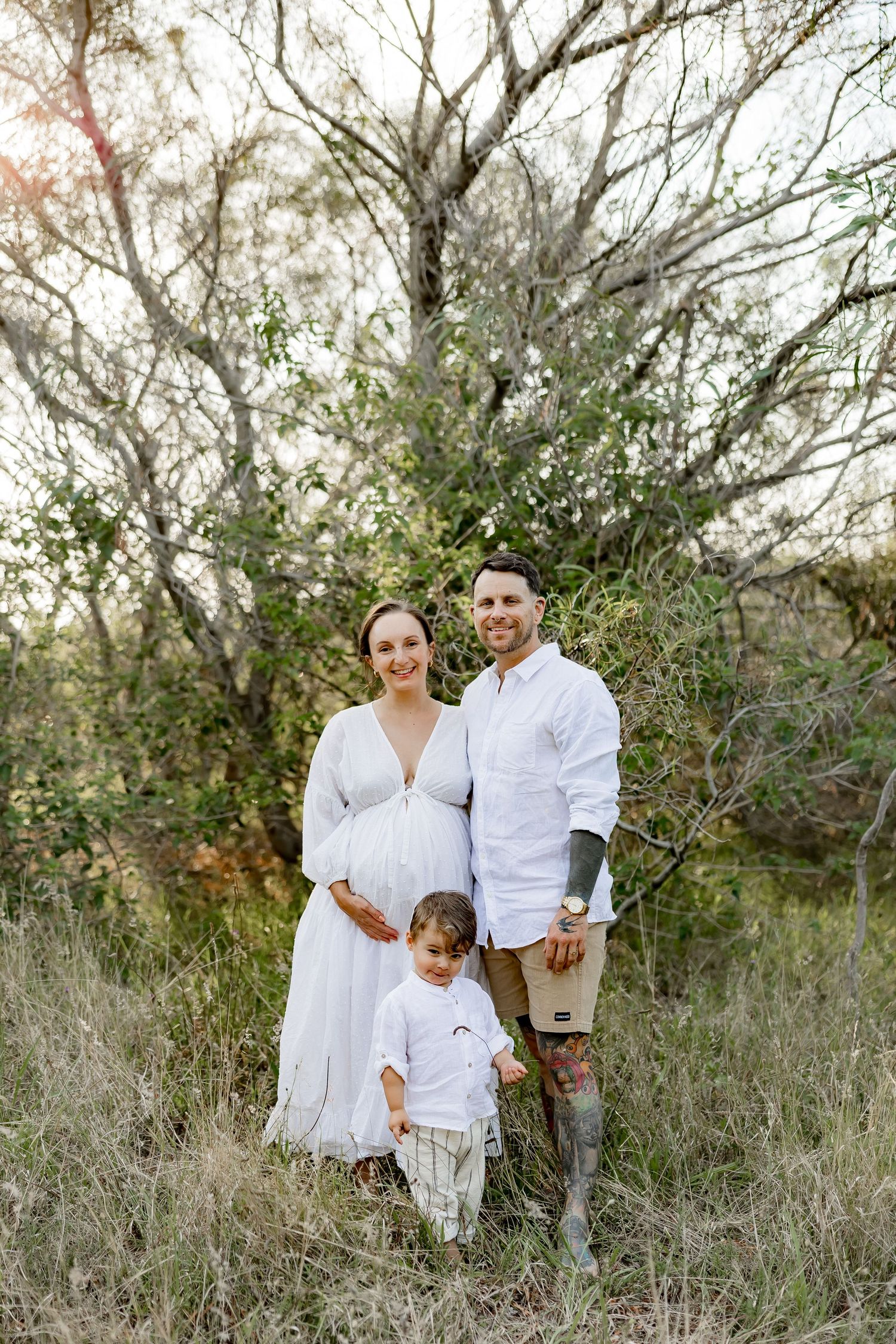 A family poses together in a natural outdoor setting wearing coordinated white outfits among tall grass and trees.
