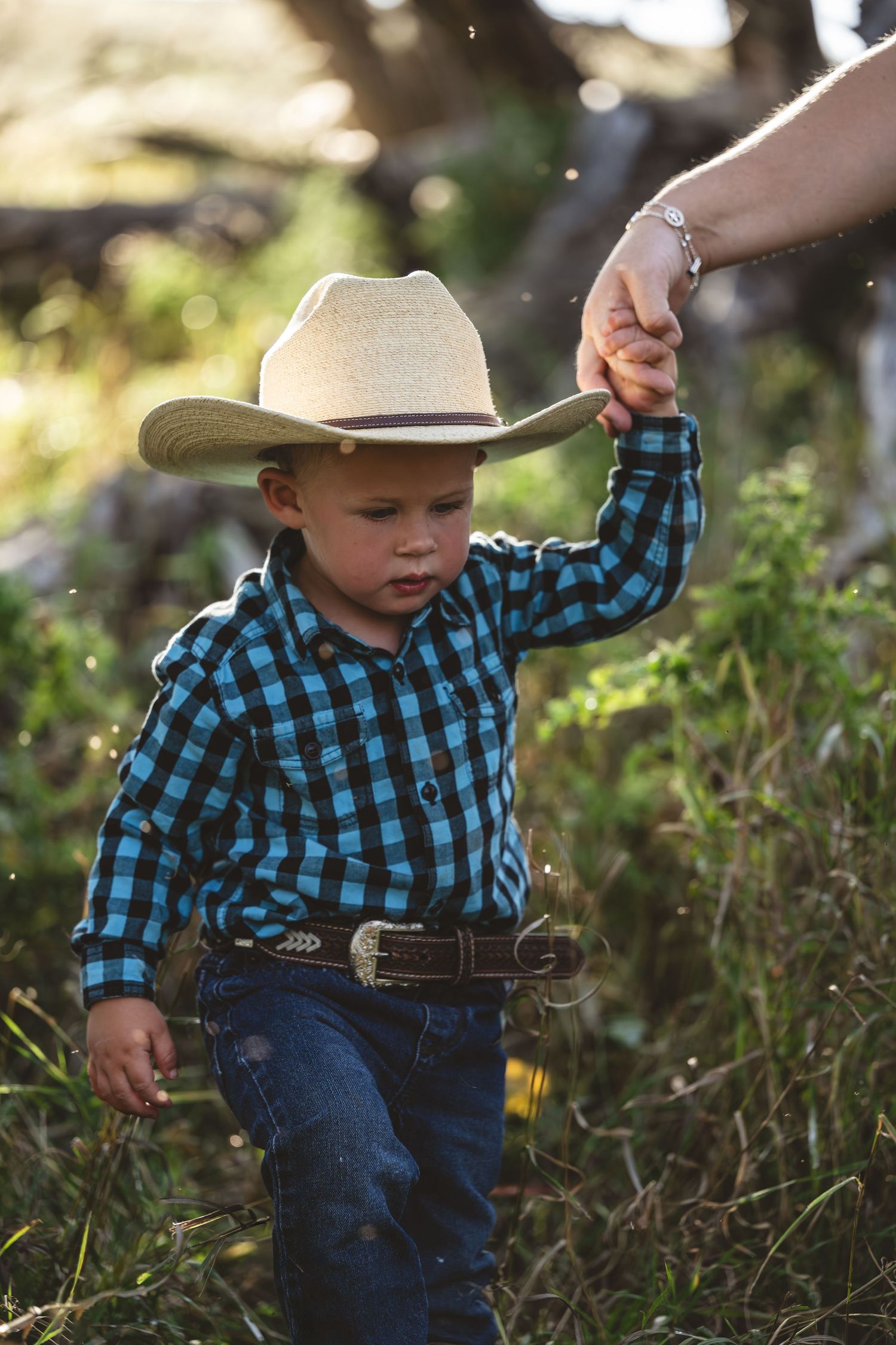 Small child in blue plaid shirt and cowboy hat walks through tall grass while being guided by adult's hand.