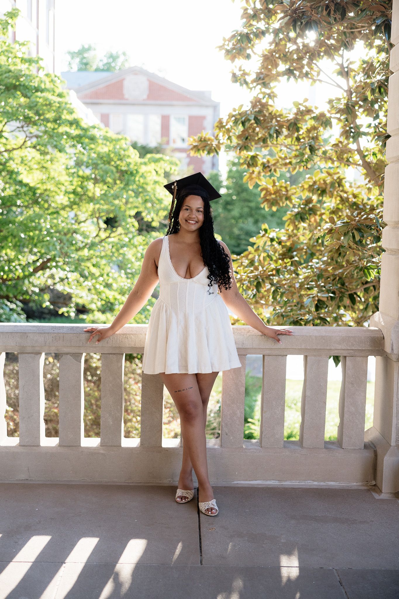 Smiling graduate in white dress and cap posing on a sunlit stone balcony.