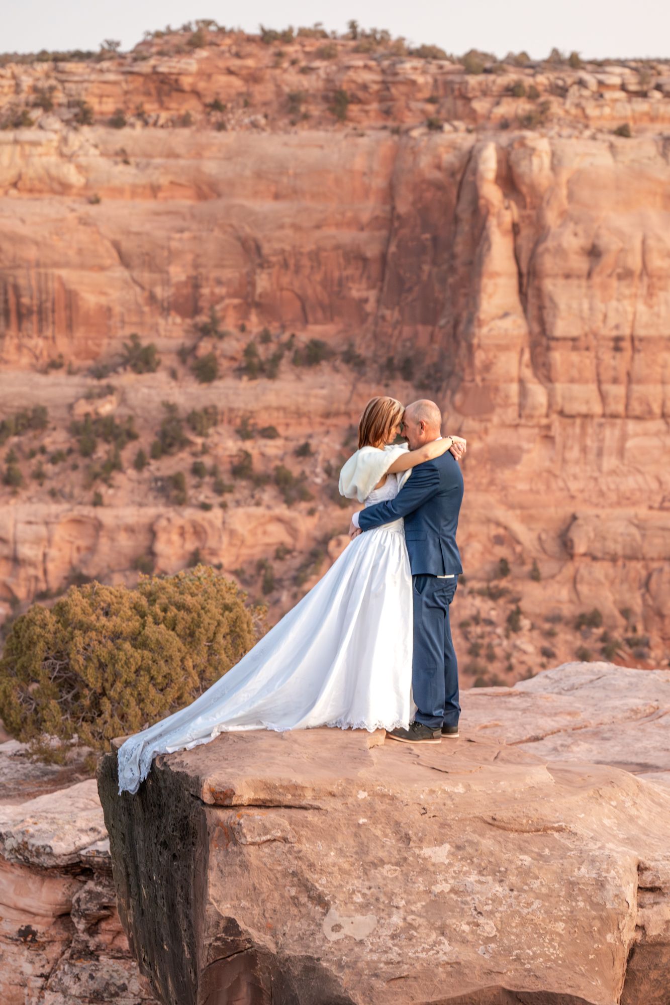 A wedding couple embraces on the edge of a red rock canyon overlook with dramatic cliffs in the background near Grand Junction.