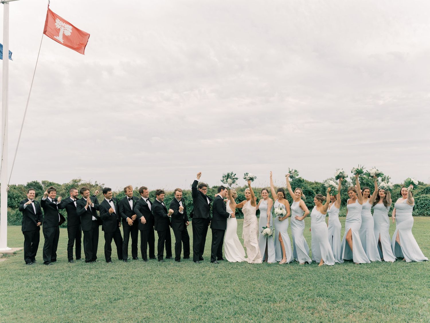 Wedding party in black and light blue attire stands in line formation on green grass field with cloudy sky.