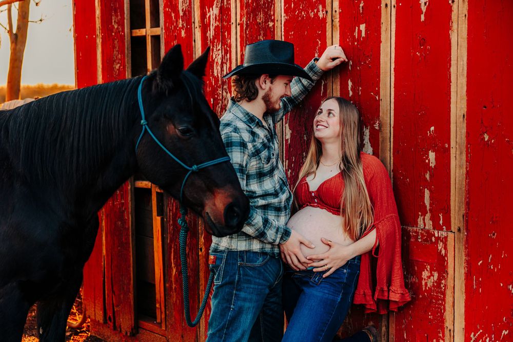 Couple embraces in front of vibrant red barn wall during rustic outdoor photoshoot.