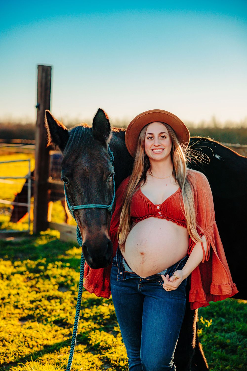 A pregnant woman in a red top and brown hat stands next to a black horse in a sunlit pasture.