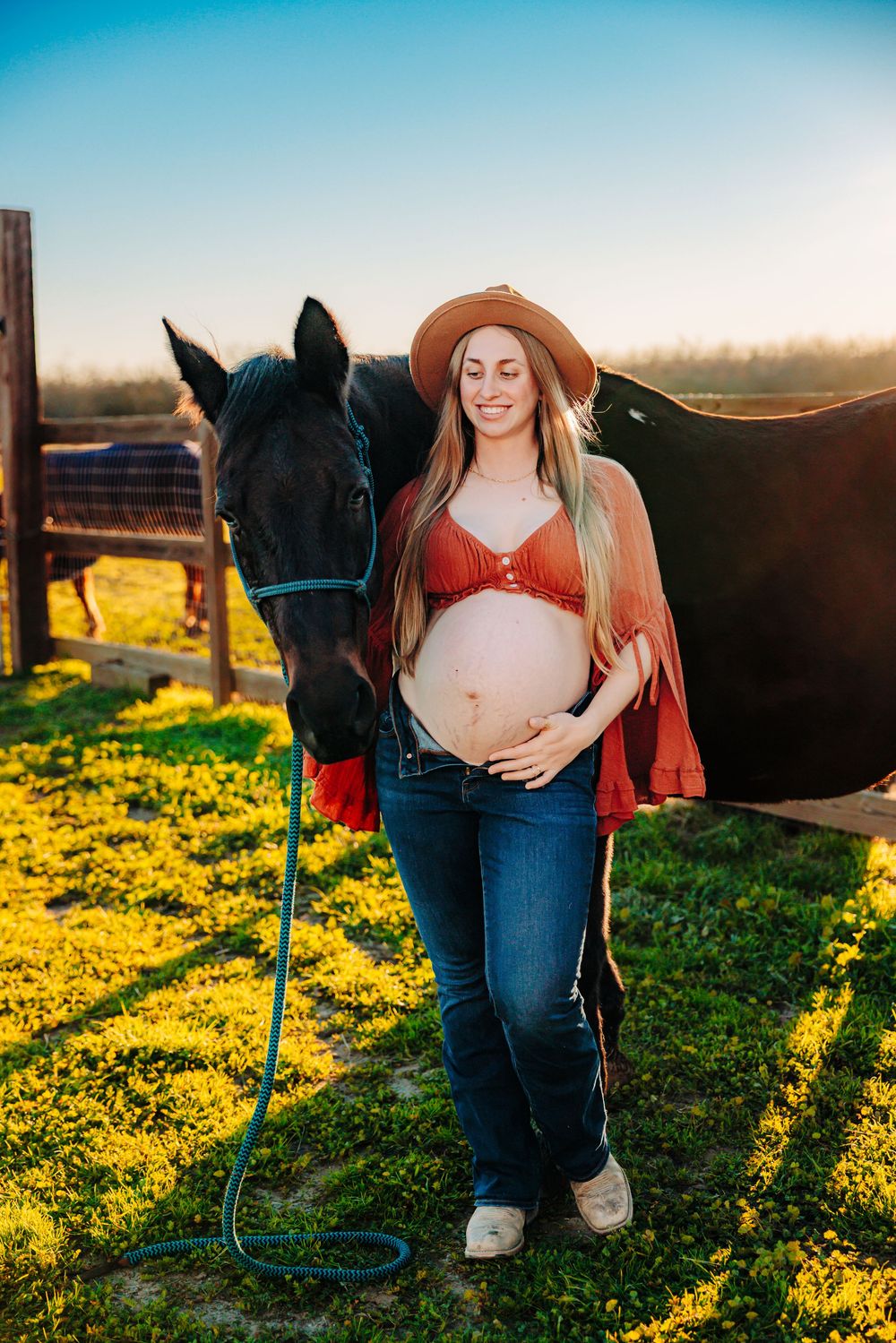 A maternity photo at sunset shows an expectant mother in a brown hat and red top standing next to a black horse in a field.
