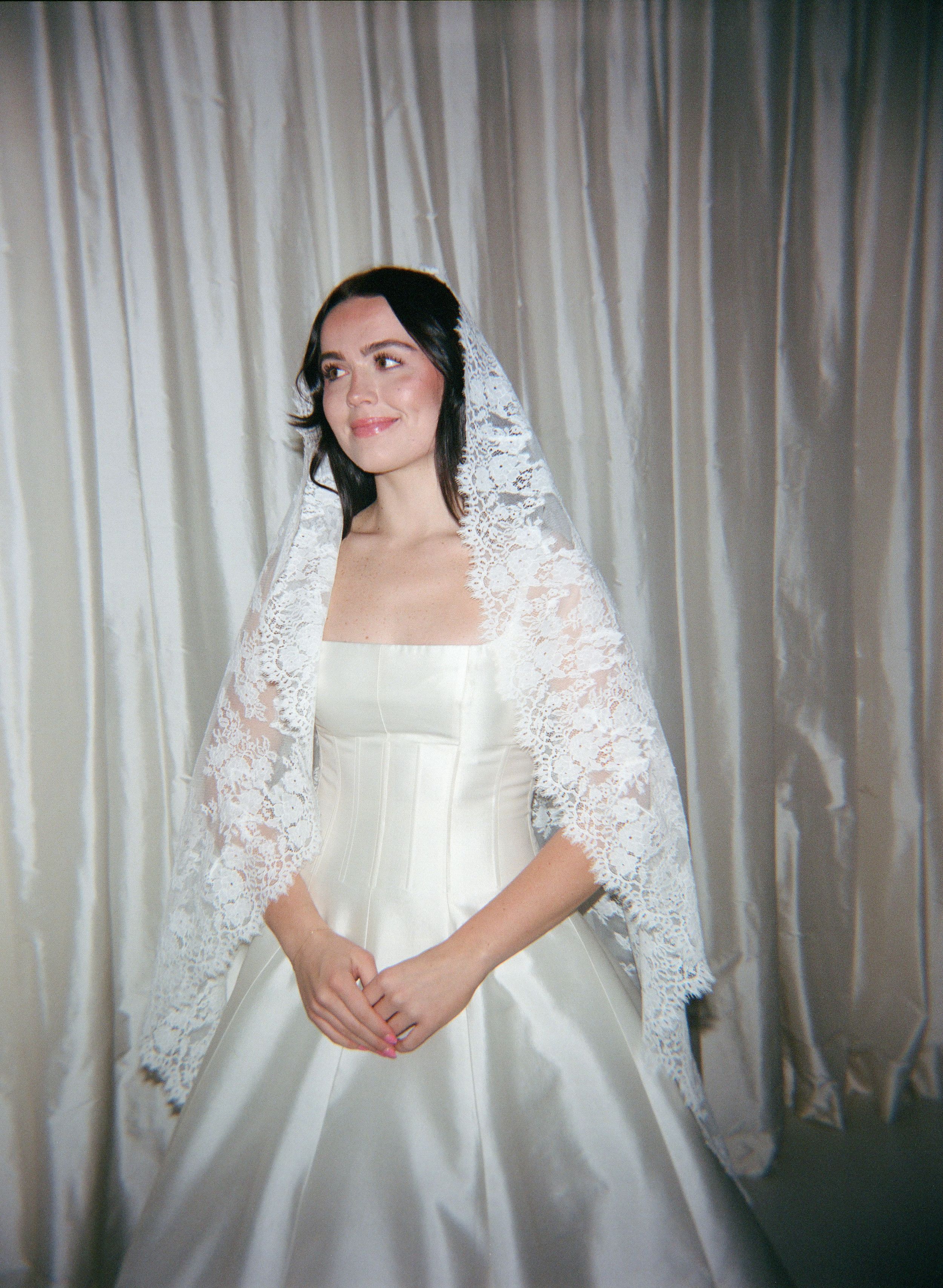 A bride wearing a traditional white wedding dress and delicate lace mantilla veil poses against a light curtain backdrop.