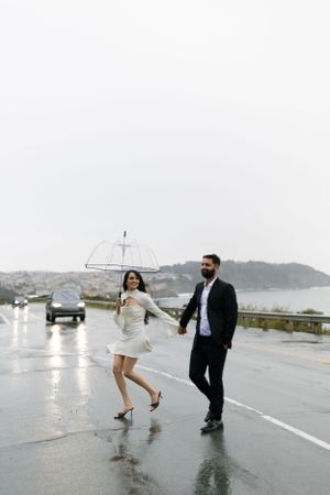 A couple walking in the rain with a clear umbrella on a coastal road.