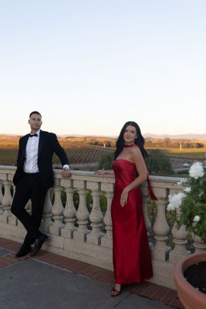 Elegant couple poses on balcony overlooking vineyard at sunset.