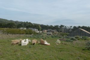 Goats graze on a grassy hillside overlooking coastal homes and buildings under a cloudy sky.