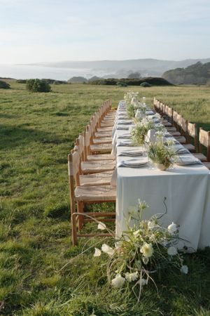 A long white dining table with rattan chairs set up outdoors in a grassy meadow overlooking coastal hills and ocean views.