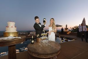 Wedding reception scene with champagne tower and cake on outdoor terrace at sunset.