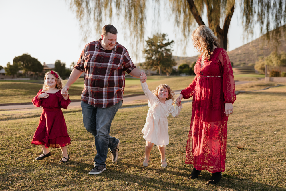 A family of four walks hand in hand across a grassy park under a willow tree at sunset.