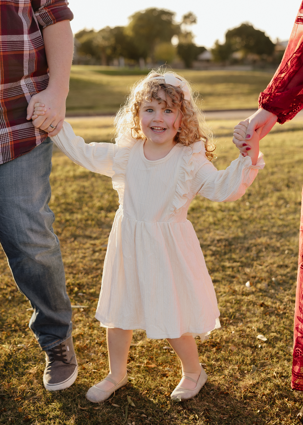 A smiling curly-haired girl in a white dress holds both parents hands while standing on grass.